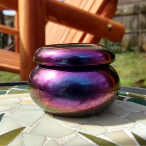 May include: A small, iridescent jar with a lid, displaying a blend of purple, blue, and pink hues. The jar sits on a mosaic tile surface, with a wooden chair in the background. The jar's colors shift depending on the light.
