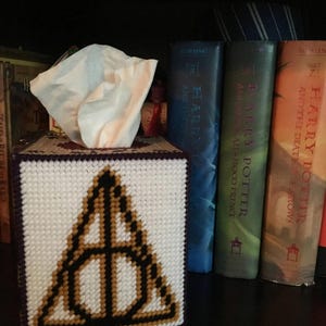 May include: A white tissue box cover with a purple border and a black and gold embroidered Deathly Hallows symbol. The tissue box cover is sitting on a shelf with a stack of Harry Potter books behind it.
