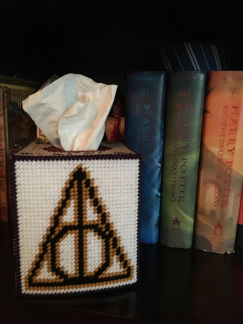 May include: A white tissue box cover with a purple border and a black and gold embroidered Deathly Hallows symbol. The tissue box cover is sitting on a shelf with a stack of Harry Potter books behind it.