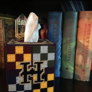 May include: A tissue box cover with a checkered pattern in purple, yellow, white, and black. The cover features a large letter "H" in the center. The tissue box cover is sitting on a shelf with several Harry Potter books behind it.