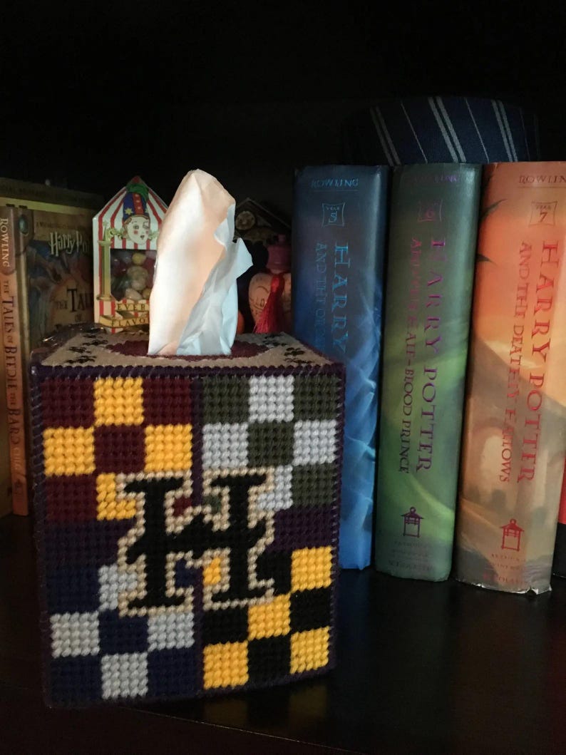 May include: A tissue box cover with a checkered pattern in purple, yellow, white, and black. The cover features a large letter "H" in the center. The tissue box cover is sitting on a shelf with several Harry Potter books behind it.