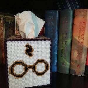 May include: A white tissue box cover with a purple border and a cross-stitch design of Harry Potter's glasses and the Deathly Hallows symbol. The tissue box cover is sitting on a shelf with a row of Harry Potter books behind it.