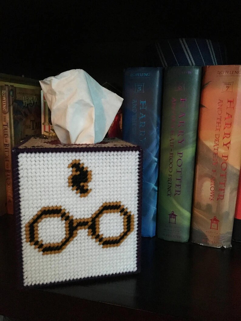 May include: A white tissue box cover with a purple border and a cross-stitch design of Harry Potter's glasses and the Deathly Hallows symbol. The tissue box cover is sitting on a shelf with a row of Harry Potter books behind it.