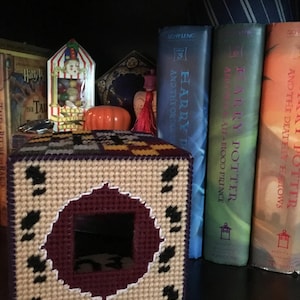 May include: A beige and purple crocheted tissue box cover with a circular opening. The cover features a black and white pattern around the opening. The cover is sitting on a shelf with Harry Potter books behind it.