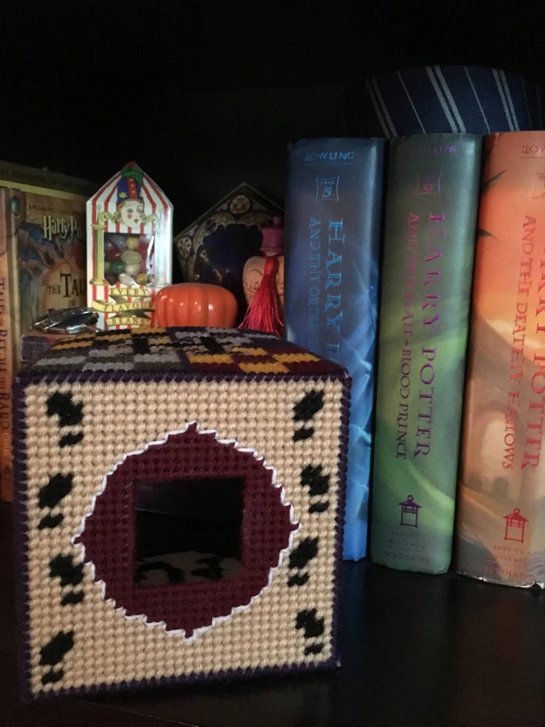 May include: A beige and purple crocheted tissue box cover with a circular opening. The cover features a black and white pattern around the opening. The cover is sitting on a shelf with Harry Potter books behind it.