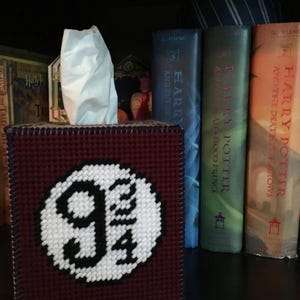 May include: A burgundy tissue box cover with a white embroidered image of the Platform 9 3/4 sign from the Harry Potter series.