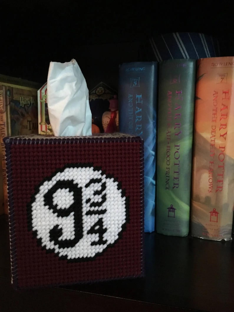 May include: A burgundy tissue box cover with a white embroidered image of the Platform 9 3/4 sign from the Harry Potter series.