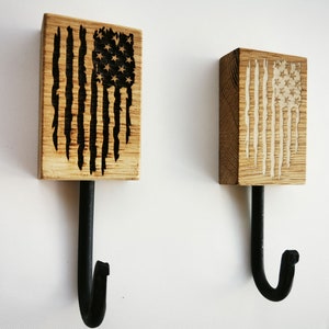 Op de afbeelding: Twee houten haken met een zwarte metalen haak die aan elk is bevestigd. Het hout is gebeitst en heeft een verweerd Amerikaans vlagontwerp dat in het hout is gebrand. Eén vlag is zwart-wit, de andere is wit-zwart.