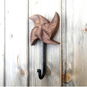 May include: A wooden wall hook shaped like a pinwheel with a black metal hook. The hook is mounted on a light wood wall.