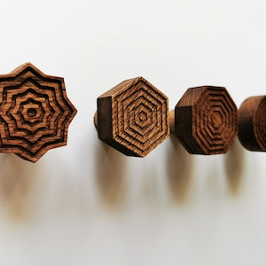 Puede incluir: Cuatro ganchos de pared de madera con diseños geométricos. Los ganchos tienen forma de estrella, hexágono, hexágono y círculo. Los ganchos son todos de madera oscura.