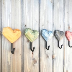 Op de afbeelding: Vijf houten hartvormige haken in verschillende kleuren, gemonteerd op een houten muur. De haken zijn geverfd in tinten geel, groen, blauw, grijs en rood.