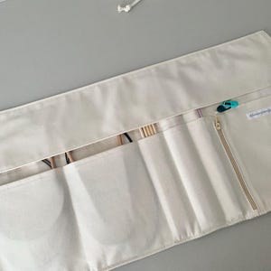 May include: A cream-coloured knitting needle case with multiple pockets and a zipped compartment. The case is open, revealing knitting needles and other tools. A small, teal and black stitch marker is attached to the zip. The case is made of a soft, fabric material.