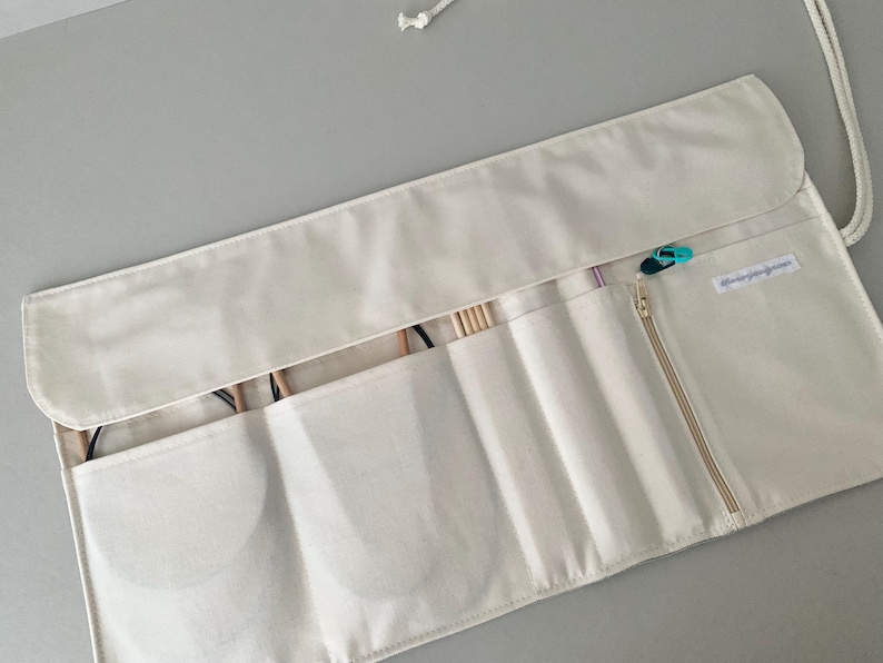 May include: A cream-coloured knitting needle case with multiple pockets and a zipped compartment. The case is open, revealing knitting needles and other tools. A small, teal and black stitch marker is attached to the zip. The case is made of a soft, fabric material.