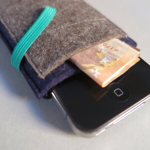 Puede incluir: Una funda para tel&eacute;fono de fieltro gris con una banda el&aacute;stica azul y verde. La funda tiene un bolsillo para una tarjeta de cr&eacute;dito o efectivo. La funda se muestra con un billete de euro en el bolsillo.