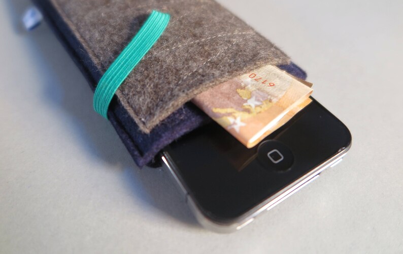 Puede incluir: Una funda para tel&eacute;fono de fieltro gris con una banda el&aacute;stica azul y verde. La funda tiene un bolsillo para una tarjeta de cr&eacute;dito o efectivo. La funda se muestra con un billete de euro en el bolsillo.