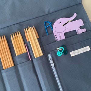 Puede incluir: Un estuche gris de tela para agujas de tejer con varias herramientas de tejido. El estuche contiene agujas de tejer de madera, un ganchillo rosa, un medidor con forma de elefante morado y marcadores de punto. Tambi&eacute;n se ve un clip azul.