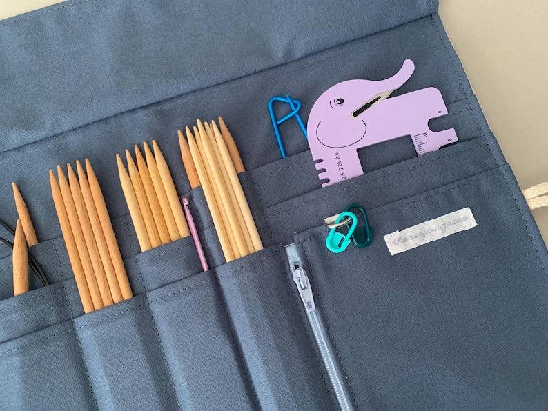 Puede incluir: Un estuche gris de tela para agujas de tejer con varias herramientas de tejido. El estuche contiene agujas de tejer de madera, un ganchillo rosa, un medidor con forma de elefante morado y marcadores de punto. Tambi&eacute;n se ve un clip azul.