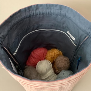Pode incluir: Vista interior de uma bolsa de m&atilde;o de fio listrada em rosa e branco. O interior &eacute; forrado com tecido azul e cont&eacute;m novelos de l&atilde; de v&aacute;rias cores. Um z&iacute;per e bolsos s&atilde;o vis&iacute;veis. Agulhas de tric&ocirc; e um marcador de ponto tamb&eacute;m est&atilde;o dentro.