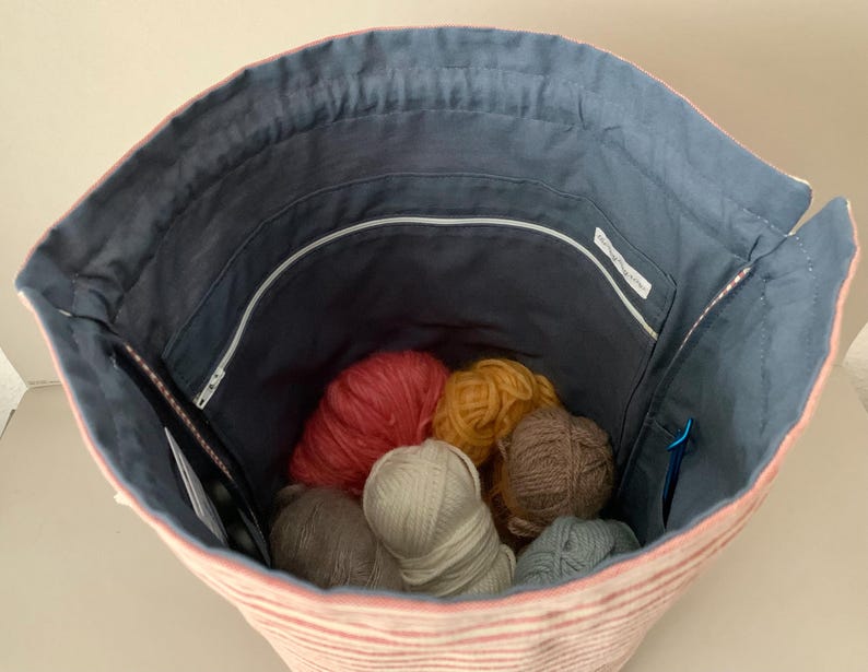 Pode incluir: Vista interior de uma bolsa de m&atilde;o de fio listrada em rosa e branco. O interior &eacute; forrado com tecido azul e cont&eacute;m novelos de l&atilde; de v&aacute;rias cores. Um z&iacute;per e bolsos s&atilde;o vis&iacute;veis. Agulhas de tric&ocirc; e um marcador de ponto tamb&eacute;m est&atilde;o dentro.