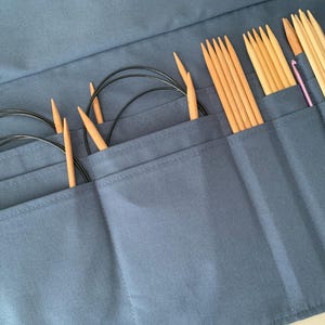 Puede incluir: Un estuche azul de tela para agujas de tejer, abierto para mostrar varias agujas. El estuche contiene varios juegos de agujas circulares con cables negros y puntas de madera, adem&aacute;s de agujas rectas de madera y un ganchillo.