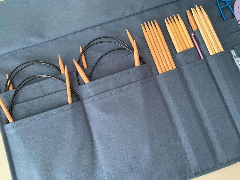 Puede incluir: Un estuche azul de tela para agujas de tejer, abierto para mostrar varias agujas. El estuche contiene varios juegos de agujas circulares con cables negros y puntas de madera, adem&aacute;s de agujas rectas de madera y un ganchillo.
