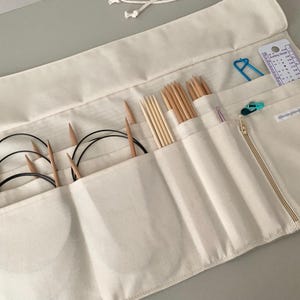 May include: Cream-coloured knitting needle organiser roll with various wooden knitting needles, circular needles, and accessories. The roll has multiple pockets and a zipped compartment. A blue stitch marker and a gauge ruler are visible.