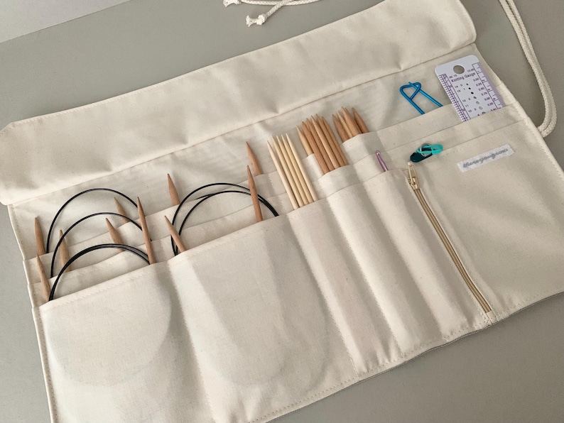 May include: Cream-coloured knitting needle organiser roll with various wooden knitting needles, circular needles, and accessories. The roll has multiple pockets and a zipped compartment. A blue stitch marker and a gauge ruler are visible.