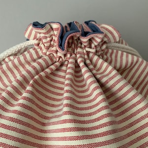 Pode incluir: Close-up de uma bolsa com cord&atilde;o com listras horizontais vermelhas e brancas. A borda superior &eacute; franzida e adornada com um tecido azul. Um cord&atilde;o branco &eacute; vis&iacute;vel.