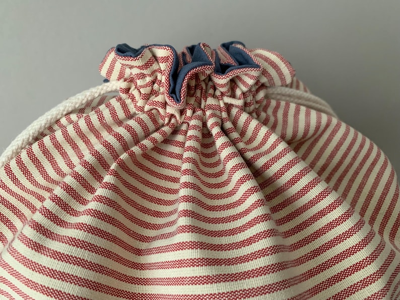 Pode incluir: Close-up de uma bolsa com cord&atilde;o com listras horizontais vermelhas e brancas. A borda superior &eacute; franzida e adornada com um tecido azul. Um cord&atilde;o branco &eacute; vis&iacute;vel.