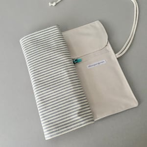 May include: A beige canvas pencil case with a grey and white striped fabric roll. The case has a flap closure and a white cord. A small, turquoise clasp is visible. The fabric roll is partially unrolled.
