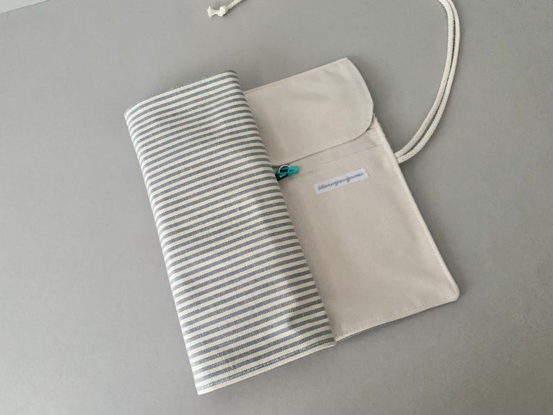May include: A beige canvas pencil case with a grey and white striped fabric roll. The case has a flap closure and a white cord. A small, turquoise clasp is visible. The fabric roll is partially unrolled.