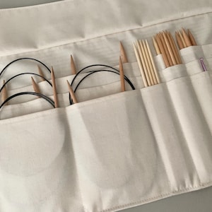May include: Cream canvas knitting needle organiser with multiple pockets. The organiser holds various wooden knitting needles, circular needles, and other knitting accessories. A zip and a few metal clips are visible.