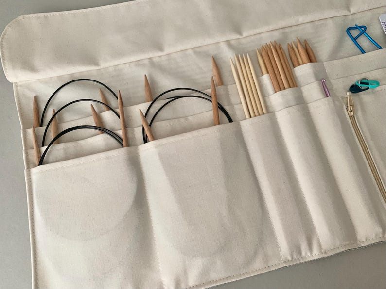 May include: Cream canvas knitting needle organiser with multiple pockets. The organiser holds various wooden knitting needles, circular needles, and other knitting accessories. A zip and a few metal clips are visible.