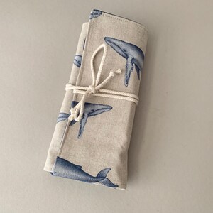 Puede incluir: Un estuche de tela beige enrollado con un patr&oacute;n repetido de ballenas azules. El estuche est&aacute; asegurado con una cuerda de color natural. Las ilustraciones de ballenas son detalladas y le dan un tema n&aacute;utico.