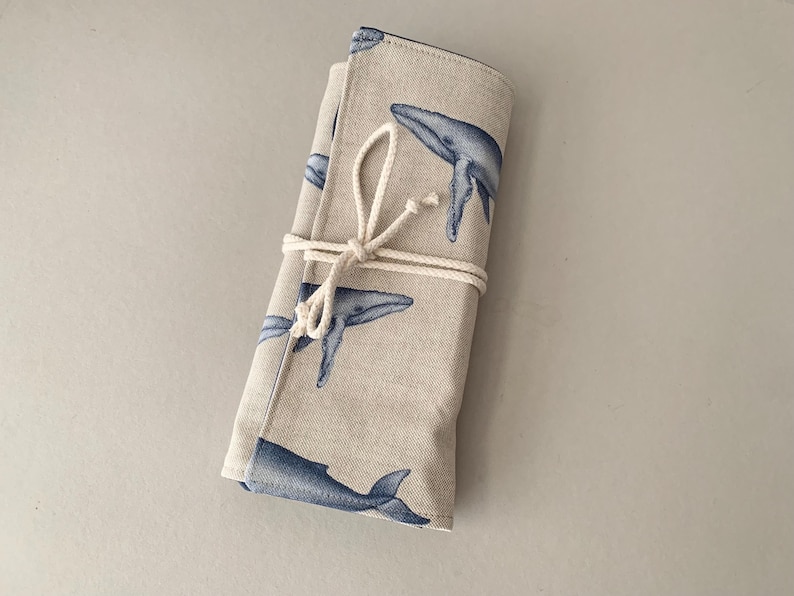 Puede incluir: Un estuche de tela beige enrollado con un patr&oacute;n repetido de ballenas azules. El estuche est&aacute; asegurado con una cuerda de color natural. Las ilustraciones de ballenas son detalladas y le dan un tema n&aacute;utico.