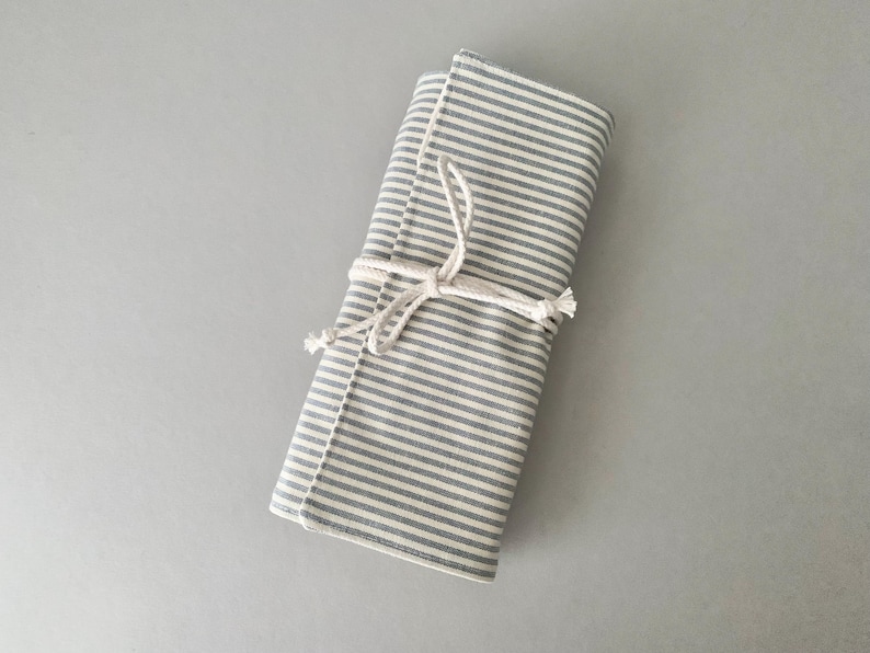 May include: A rolled fabric case with grey and white stripes, tied with a natural rope. The case is made of a soft material, possibly for storage. The design is simple and minimalist, with a focus on natural materials.