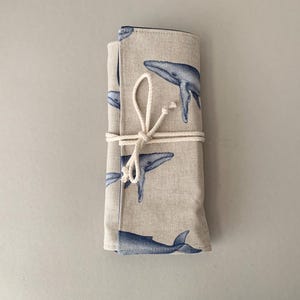Puede incluir: Un estuche de tela beige enrollado con un patr&oacute;n repetido de ballenas azules. El estuche est&aacute; asegurado con una cuerda color crema atada con un lazo. Las ilustraciones de ballenas son detalladas y art&iacute;sticas.