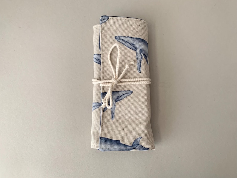 Puede incluir: Un estuche de tela beige enrollado con un patr&oacute;n repetido de ballenas azules. El estuche est&aacute; asegurado con una cuerda color crema atada con un lazo. Las ilustraciones de ballenas son detalladas y art&iacute;sticas.