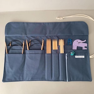 Puede incluir: Un estuche enrollable para agujas de tejer de tela azul gris&aacute;ceo con varias agujas de tejer, agujas circulares y accesorios. El estuche tiene m&uacute;ltiples bolsillos, un cierre de cremallera y un cierre de cord&oacute;n. Un marcador de puntada con forma de elefante morado y otros accesorios son visibles.