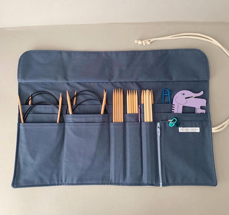 Puede incluir: Un estuche enrollable para agujas de tejer de tela azul gris&aacute;ceo con varias agujas de tejer, agujas circulares y accesorios. El estuche tiene m&uacute;ltiples bolsillos, un cierre de cremallera y un cierre de cord&oacute;n. Un marcador de puntada con forma de elefante morado y otros accesorios son visibles.