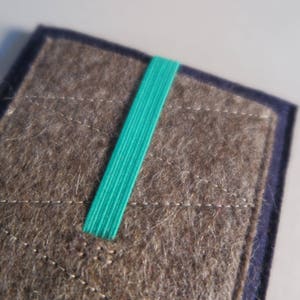 Puede incluir: Una funda de fieltro marr&oacute;n con una banda el&aacute;stica turquesa. La funda tiene un patr&oacute;n cosido y un borde azul oscuro.