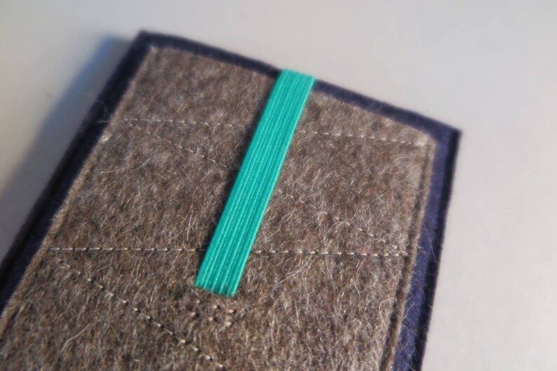 Puede incluir: Una funda de fieltro marr&oacute;n con una banda el&aacute;stica turquesa. La funda tiene un patr&oacute;n cosido y un borde azul oscuro.