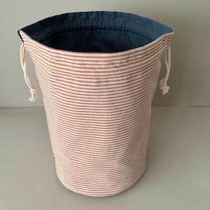 Pode incluir: Um saco de armazenamento cil&iacute;ndrico de tecido com exterior listrado em vermelho e branco e interior azul escuro. A bolsa tem um fecho de cord&atilde;o com cord&otilde;es brancos. A bolsa tem aproximadamente 25 cm de altura e 20 cm de di&acirc;metro.