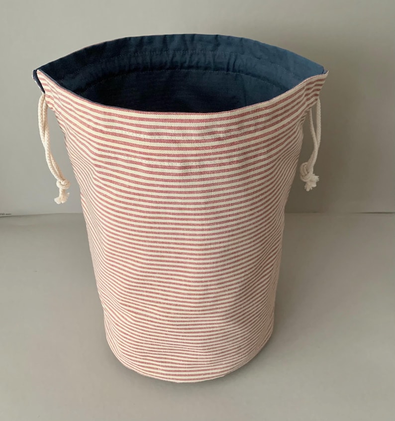 Pode incluir: Um saco de armazenamento cil&iacute;ndrico de tecido com exterior listrado em vermelho e branco e interior azul escuro. A bolsa tem um fecho de cord&atilde;o com cord&otilde;es brancos. A bolsa tem aproximadamente 25 cm de altura e 20 cm de di&acirc;metro.