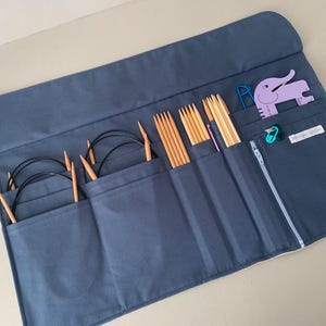 Puede incluir: Un estuche organizador de agujas de tejer azul marino con m&uacute;ltiples bolsillos. Los bolsillos contienen varias agujas de tejer de madera, agujas circulares y otros accesorios de tejido. Tambi&eacute;n se ve una herramienta morada con forma de elefante y un marcador de puntada turquesa.