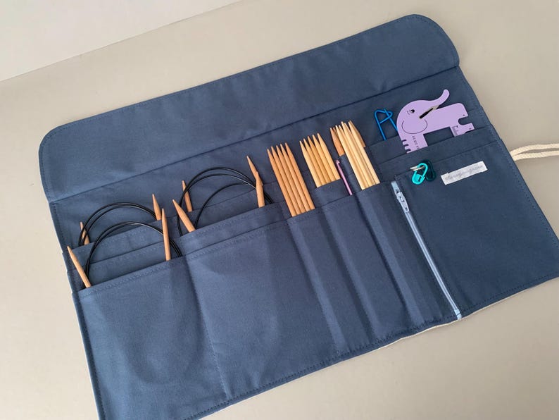 Puede incluir: Un estuche organizador de agujas de tejer azul marino con m&uacute;ltiples bolsillos. Los bolsillos contienen varias agujas de tejer de madera, agujas circulares y otros accesorios de tejido. Tambi&eacute;n se ve una herramienta morada con forma de elefante y un marcador de puntada turquesa.