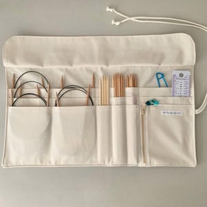 May include: Cream canvas knitting needle organiser with multiple pockets and a zip closure. The organiser holds various knitting needles, circular needles, stitch markers, and a knitting gauge. A white drawstring is attached for closure.