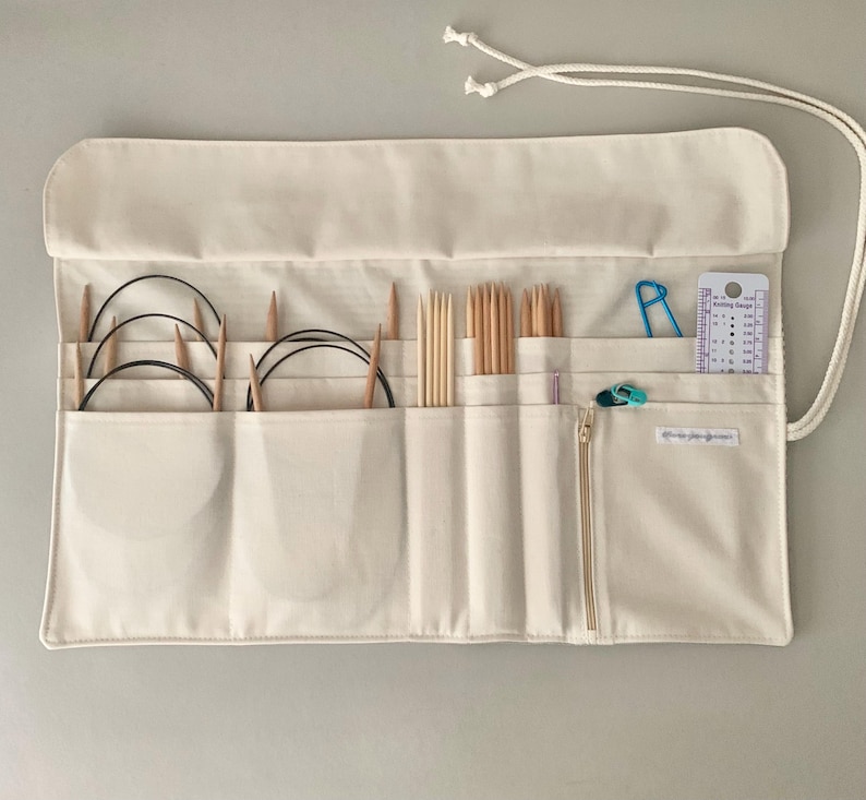 May include: Cream canvas knitting needle organiser with multiple pockets and a zip closure. The organiser holds various knitting needles, circular needles, stitch markers, and a knitting gauge. A white drawstring is attached for closure.