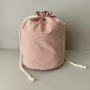 Pode incluir: Uma bolsa cil&iacute;ndrica com cord&atilde;o com um padr&atilde;o de listras vermelhas e brancas. A bolsa &eacute; feita de tecido e tem um fecho de cord&atilde;o branco. A parte superior da bolsa &eacute; franzida e tem um forro azul.