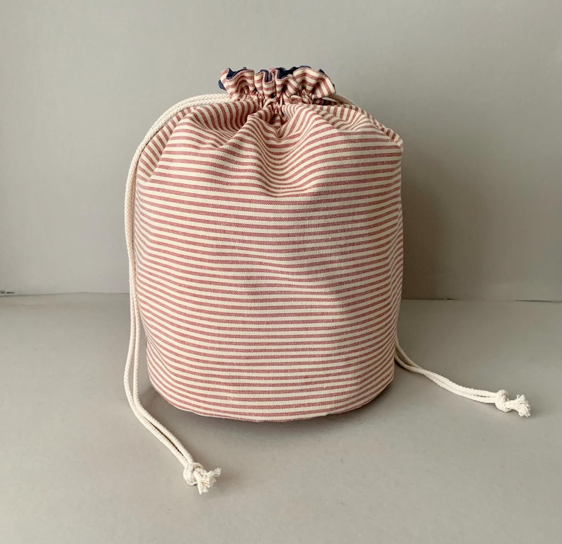 Pode incluir: Uma bolsa cil&iacute;ndrica com cord&atilde;o com um padr&atilde;o de listras vermelhas e brancas. A bolsa &eacute; feita de tecido e tem um fecho de cord&atilde;o branco. A parte superior da bolsa &eacute; franzida e tem um forro azul.
